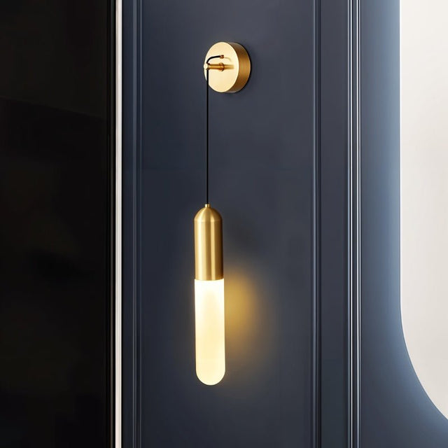 "A minimalist brass wall lamp with a long, slender cylindrical shade casting a soft, warm light on a dark blue paneled wall."
