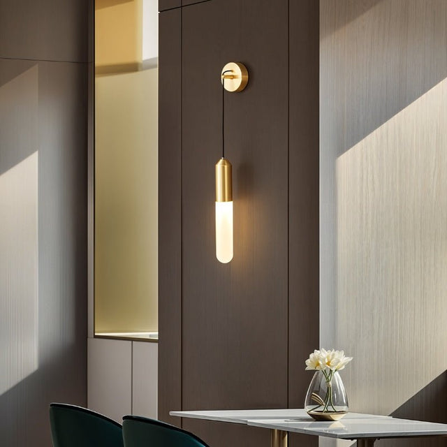 "A minimalist brass wall sconce with a long, slender cylindrical shade casting a soft glow on a dark wall in a modern living room."