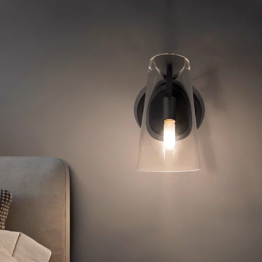 "A modern brass wall lamp with a clear acrylic shade, lit on a dark wall."
