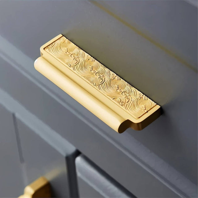 "A gold textured Erdas pull bar handle mounted on a gray cabinet."