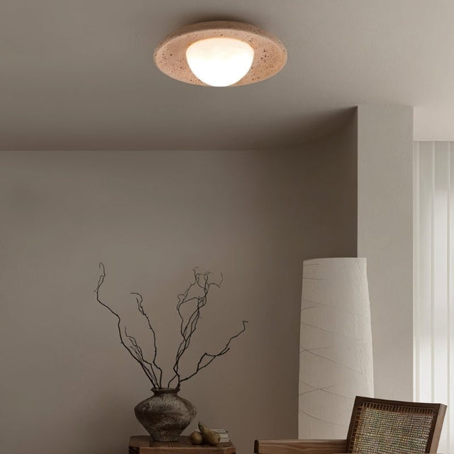"A modern, minimalist flush mount light with a round travertine base and frosted globe, mounted on a living room ceiling, providing a warm, ambient glow above a seating area with a decorative vase and chair."
