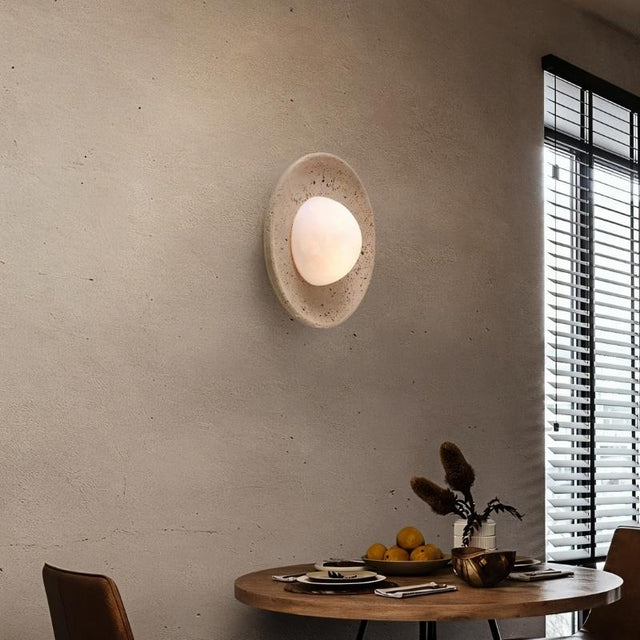 "Modern, minimalist wall light with a round travertine base and frosted globe, mounted on a textured dining room wall, providing a warm, ambient glow above a round dining table."