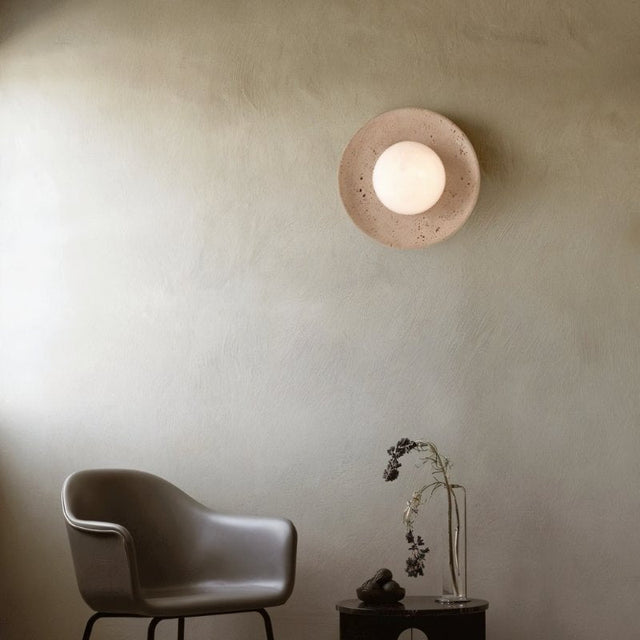 "Modern, minimalist wall mount light with round travertine base and frosted globe on a textured living room wall, providing soft ambient lighting next to a modern chair and side table with a vase."