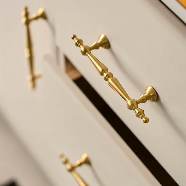 "Ornate brass pull bar handles with a classic design on light-colored drawers."