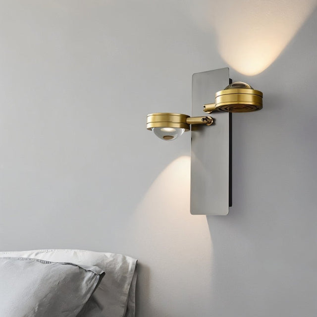 "A modern wall lamp with two adjustable lights, gunmetal finish, and glass lenses, mounted on a bedroom wall beside a pillow."