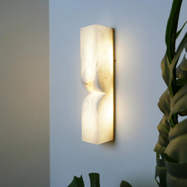 "A double curved alabaster wall lamp mounted beside a plant, creating a warm and inviting atmosphere."