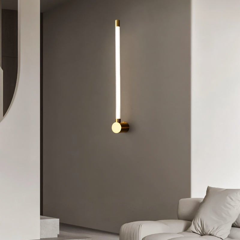 "A sleek, modern brass wall lamp with a long, thin light bar mounted vertically on a gray wall, illuminating a contemporary living room."