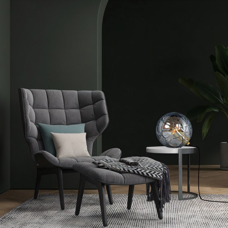 "A modern table lamp with a spherical patterned glass shade, placed on a side table in a contemporary living room with a grey armchair and dark walls."