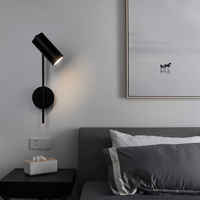 "A minimalist industrial black bedside reading lamp with a cylindrical shade and adjustable arm, mounted on a gray wall above a bed with a gray headboard and next to a black side table with a tissue box."