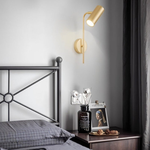 "A minimalist industrial gold bedside reading lamp with a cylindrical shade and adjustable arm, mounted on a white wall next to a bed with a black metal frame."