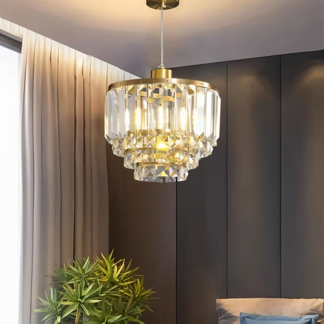 "A tiered crystal pendant light with a brass frame, hanging in a living room with a plant in the foreground."