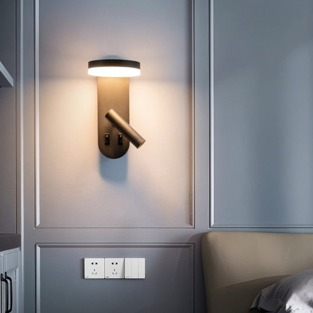 "A black wall lamp with an LED panel and a spotlight, installed above a light switch, next to a beige headboard."