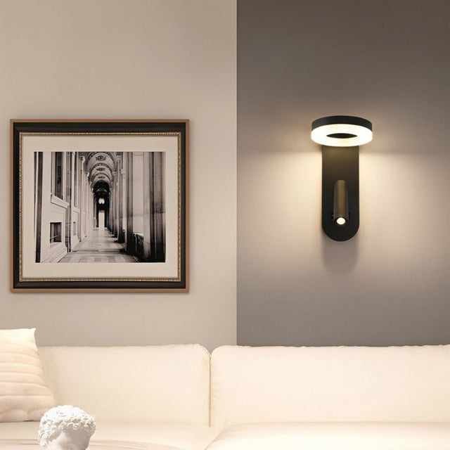 "A black wall lamp with an LED panel and a spotlight, installed in a living room with a picture frame and a white sofa."