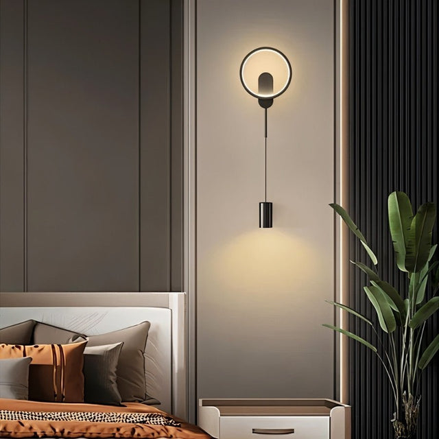"A modern black wall lamp with a circular and cylindrical design illuminates a contemporary bedroom."