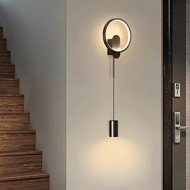 "A modern black wall lamp with a circular and cylindrical design illuminates a contemporary staircase."