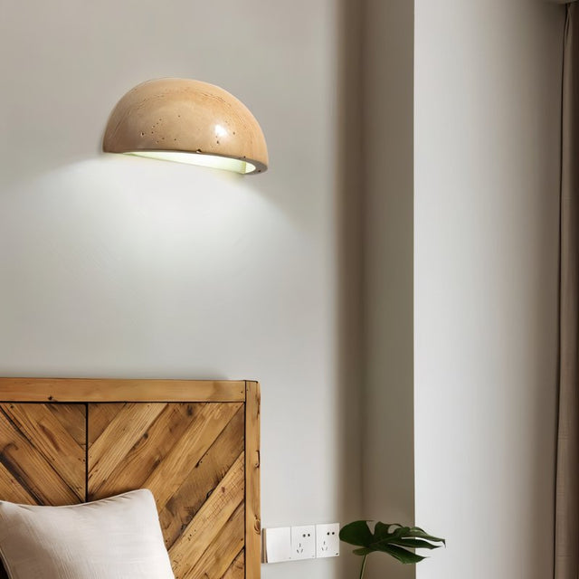 "A modern, semi-circular wall lamp made of yellow travertine, mounted on a wall above a wooden headboard in a minimalist bedroom, providing a soft, ambient light."