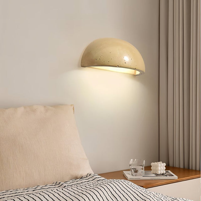 "A modern, semi-circular wall lamp made of yellow travertine, casting a warm glow in a dark bedroom, highlighting its smooth surface and organic texture."