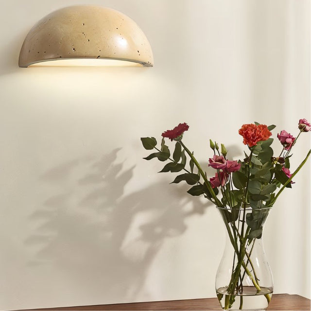 "A minimalist, semi-circular wall lamp made of yellow travertine, mounted on a plain wall, emitting a soft, ambient light, with a vase of flowers casting a shadow beside it."