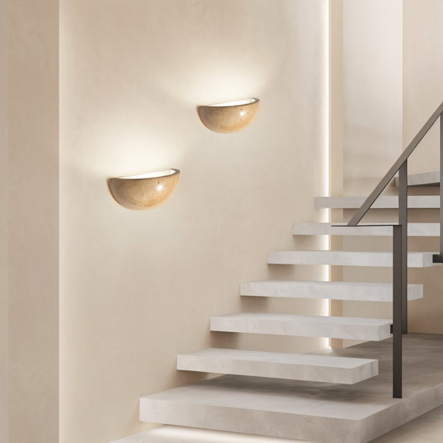 "Two modern, semi-circular wall lamps made of yellow travertine, mounted on a wall beside a stairway, providing soft, ambient lighting."