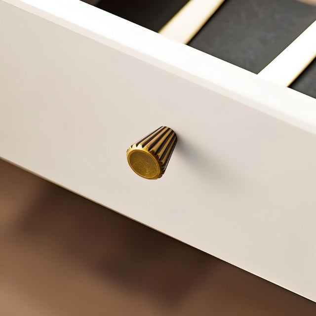 "A contemporary brass cone-shaped knob with sleek vertical grooves, installed on a white drawer, showcasing its smooth, polished circular face."