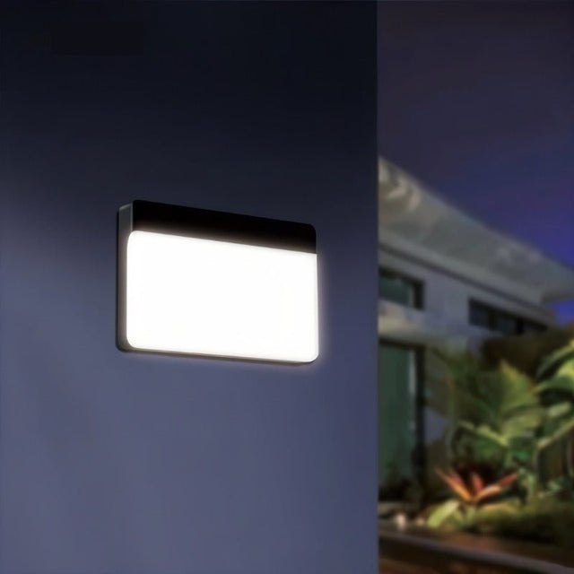 "A minimalist rectangular outdoor wall lamp, lit and mounted on a dark wall at night, providing soft illumination."