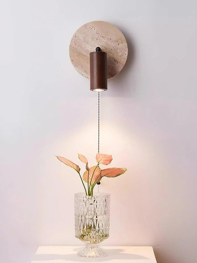 "Yellow travertine round wall lamp with wooden cylindrical shade and pull chain above a glass vase, modern reading light."