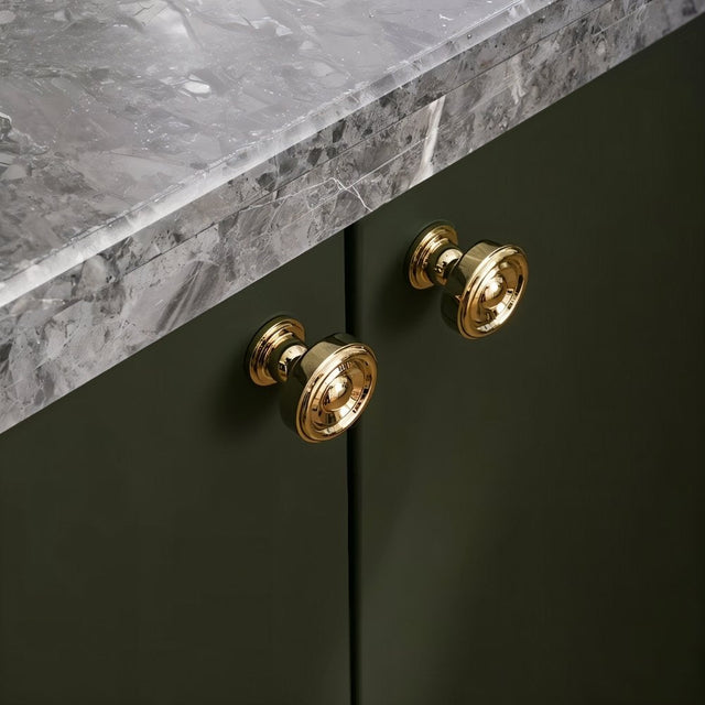 "Pair of brass knobs with a tapered spindle design and intricate finials on a green cabinet with a marble countertop."