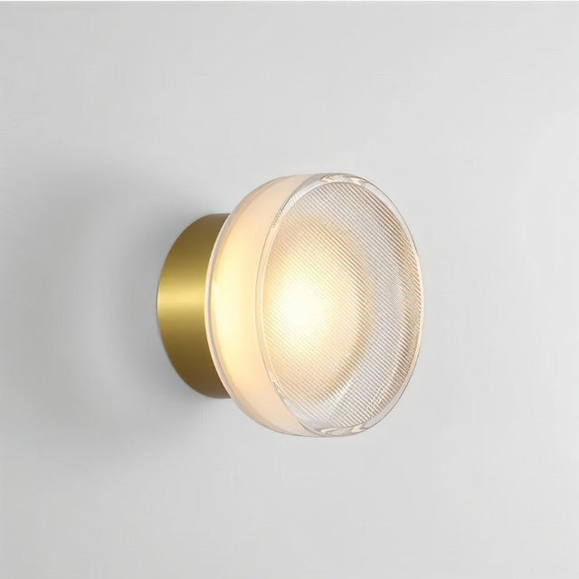 "A modern, minimalist wall lamp with a circular glass diffuser and a sleek brass frame, emitting a warm, radiant glow."