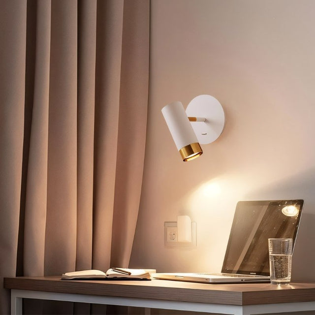 "A white wall lamp with a round base and a cylindrical shade, shown lit up on a light gray wall above a desk with a laptop, a book, and a glass of water."