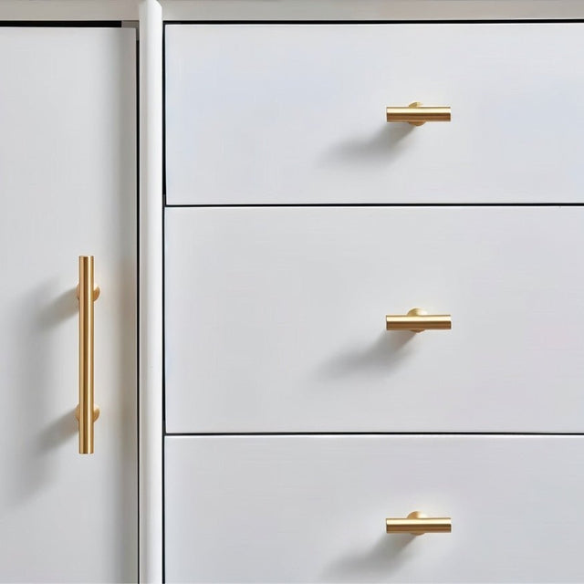 "Gold brass knobs and pull bars with cylindrical design, mounted on a white dresser with drawers and a cabinet door."