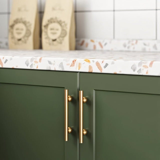 "Gold brass pull bars with cylindrical design, mounted on dark green kitchen cabinets with a terrazzo countertop."
