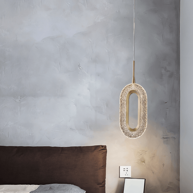 Single Luxin Pendant Light with sculptural oval LED design, hanging beside a brown bed in a minimalist bedroom with textured wall.