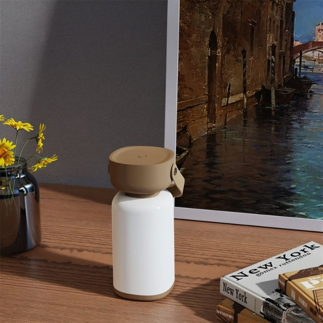 "A beige and white cylindrical table lamp with a handle, sitting on a table with books and a framed painting."