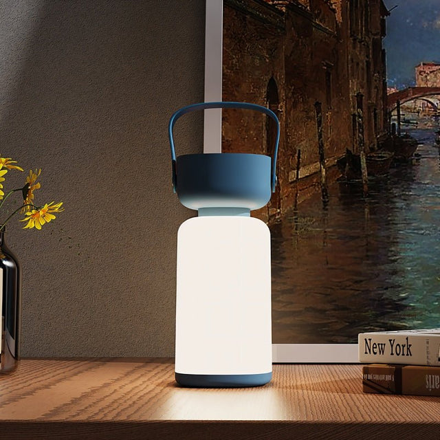 "A blue and white cylindrical table lamp with a handle, lit up with a soft glow, sitting on a wooden table with books and a framed painting."
