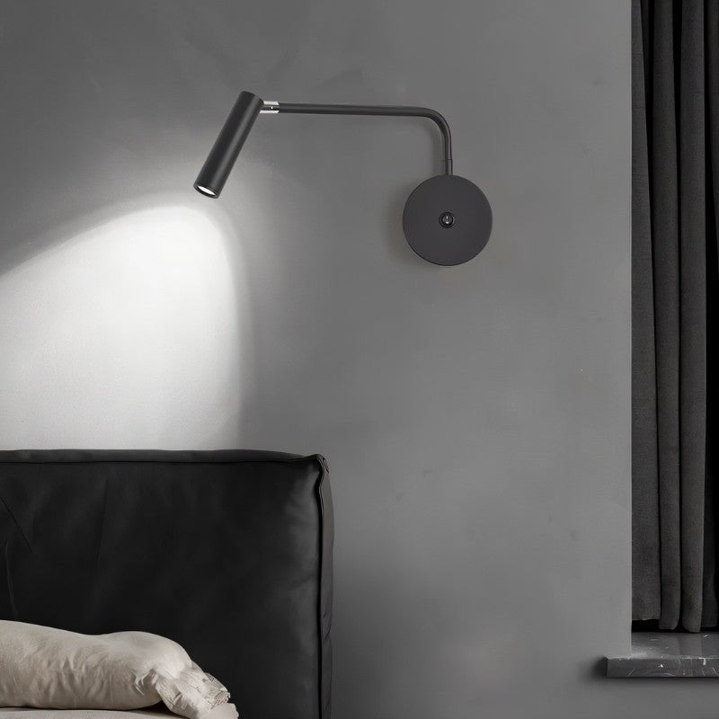 "A black wall lamp with a pivoting arm and cylindrical head, lit up on a gray wall, next to a black bed and curtains."