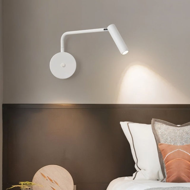 "A white wall lamp with a pivoting arm and cylindrical head, installed above a bedside table with a round object, next to a dark brown headboard."