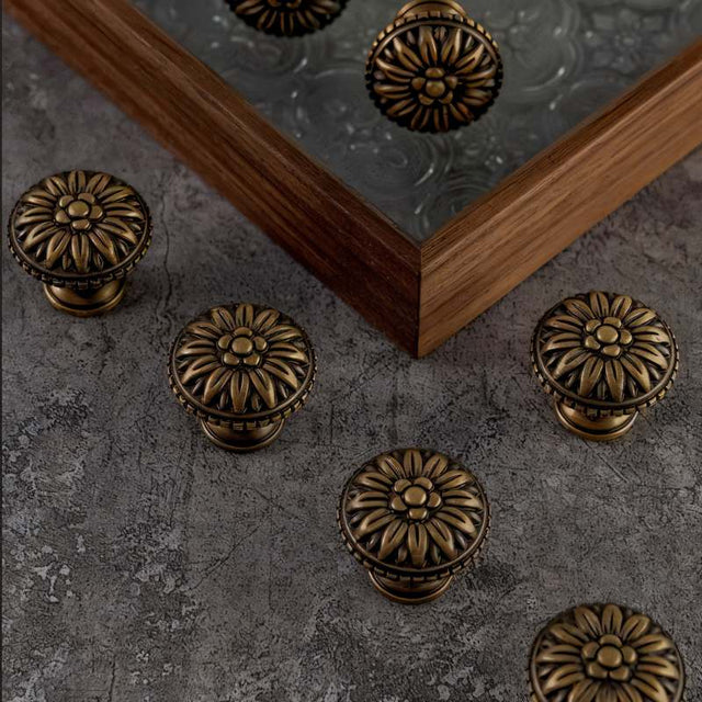 "Set of antique brass floral knobs with raised petal detailing on a textured gray surface and wooden frame."