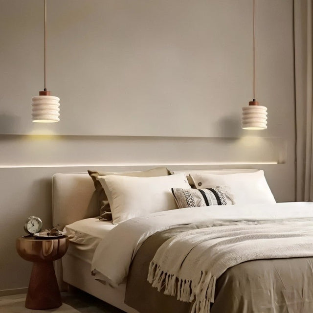 "Two travertine pendant lights with a ribbed design hanging over a bed with a warm light."