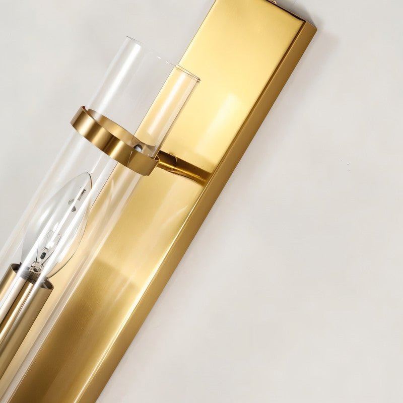 "Close-up of a modern brass wall lamp with a rectangular base and a cylindrical glass shade, showing the light bulb inside."