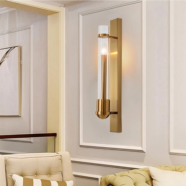 "A modern brass wall lamp with a rectangular base and a cylindrical glass shade mounted on a light-colored wall in a contemporary living room."