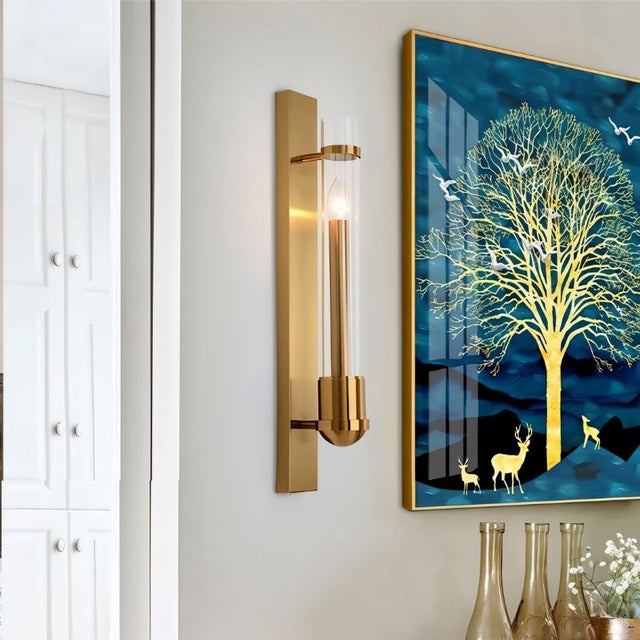 "A modern brass wall lamp with a rectangular base and a cylindrical glass shade mounted on a white wall in a living room, next to a framed painting of a tree."