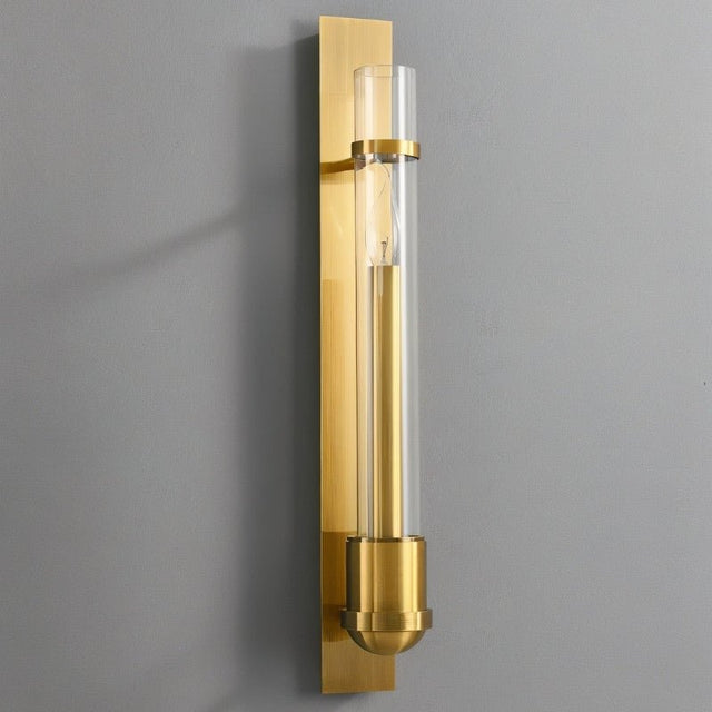 "A modern brass wall lamp with a rectangular base and a cylindrical glass shade mounted on a gray wall."