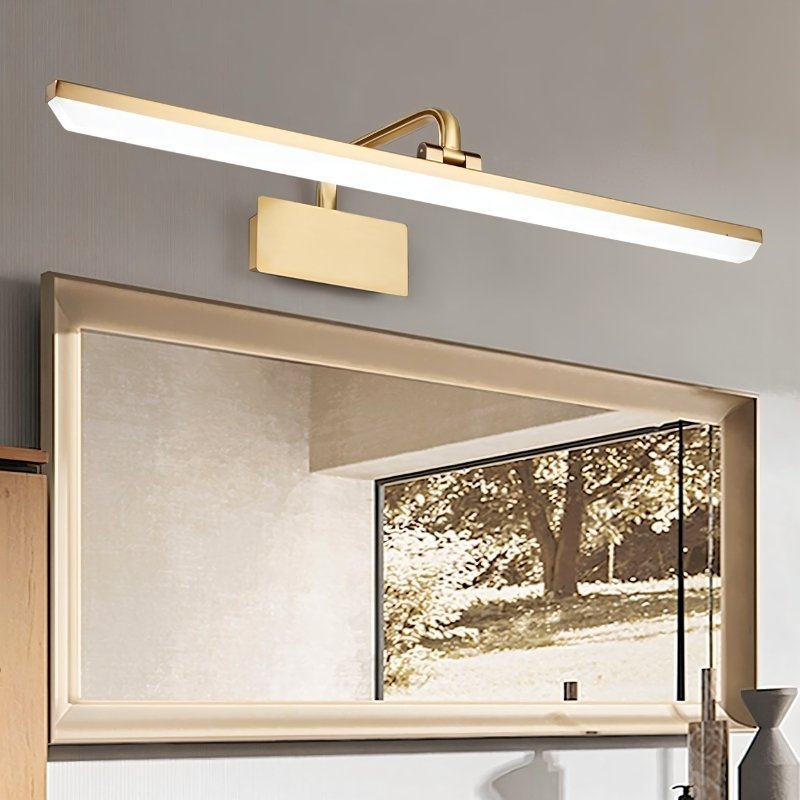 "A gold-finish LED vanity light mounted above a framed bathroom mirror with a reflection of a tree."