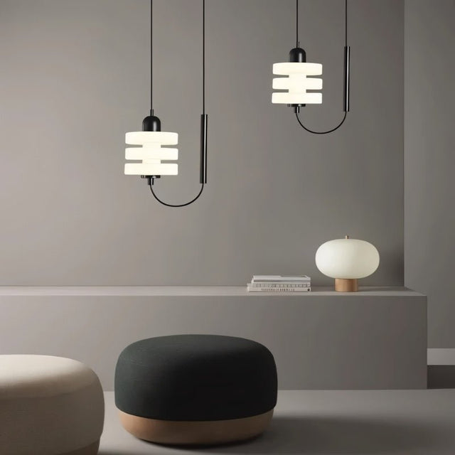 "Two modern pendant lights with layered frosted glass diffusers and black iron frames, hanging above a shelf with ottomans in the foreground."