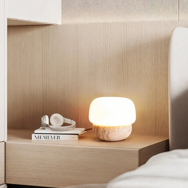 "Minimalist table lamp with a travertine base and glass shade on a wooden nightstand"