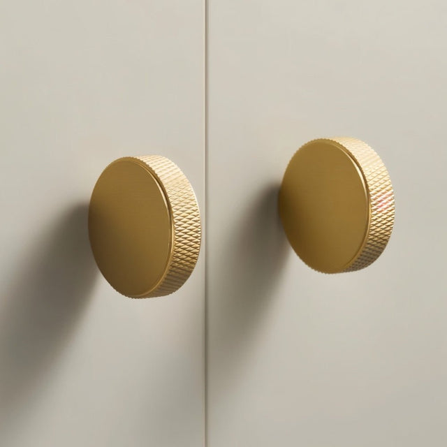 "Two gold brass cylindrical knobs with knurled texture on a light gray cabinet door."