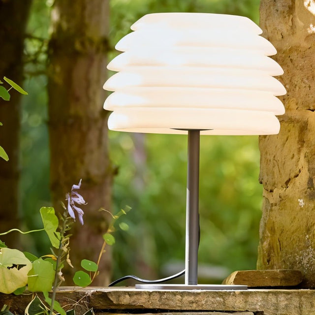 "A white table lamp with a layered, wave-like diffuser, lit up on a stone ledge in a garden with plants."
