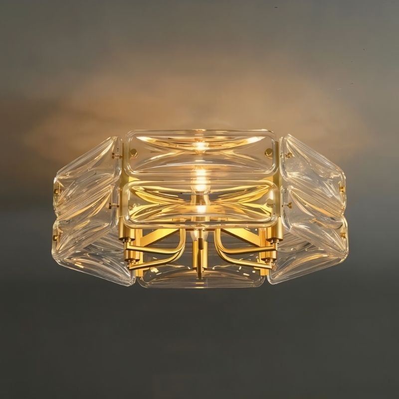 "A close-up view of a modern flush mount ceiling light with a geometric glass shade and gold frame, illuminated against a dark background."