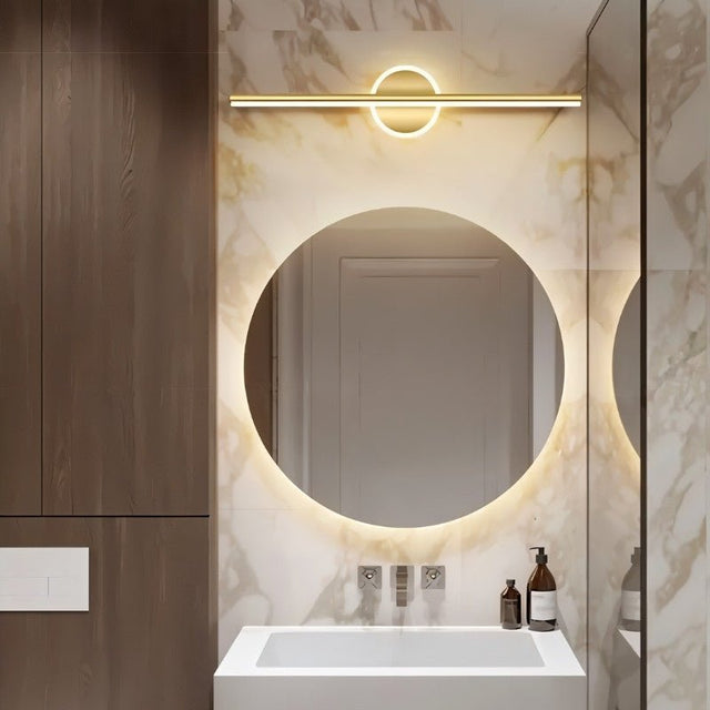 "A modern gold wall lamp with a cylindrical bar intersecting a circular base, mounted above a round mirror in a contemporary bathroom with marble walls."