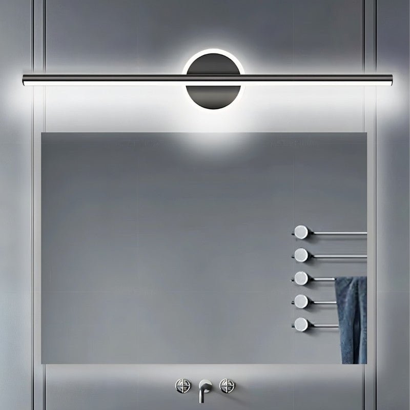 "A modern black wall lamp with a cylindrical bar intersecting a circular base, mounted above a rectangular mirror in a contemporary bathroom with a gray tiled wall and a towel rack."